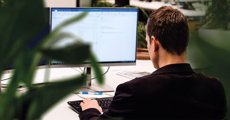 Collega van Techni Team werkt achter een beeldscherm in een kantoor met planten, gezien van achteren