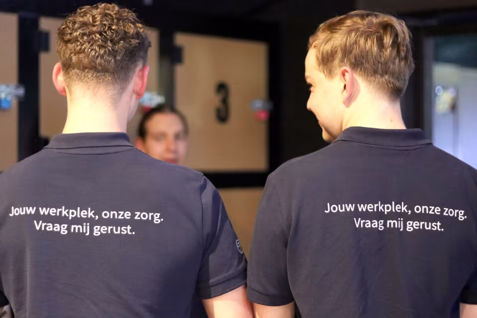 Twee collega’s gezien van achteren in poloshirts met de tekst ‘Jouw werkplek, onze zorg. Vraag mij gerust.’