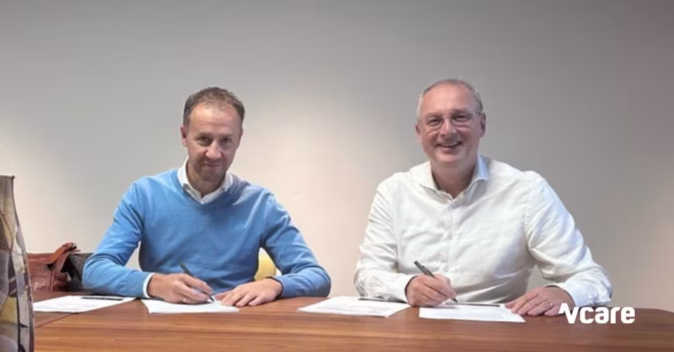 Twee mannen zitten aan een tafel met papieren, met het Vcare-logo rechtsonder