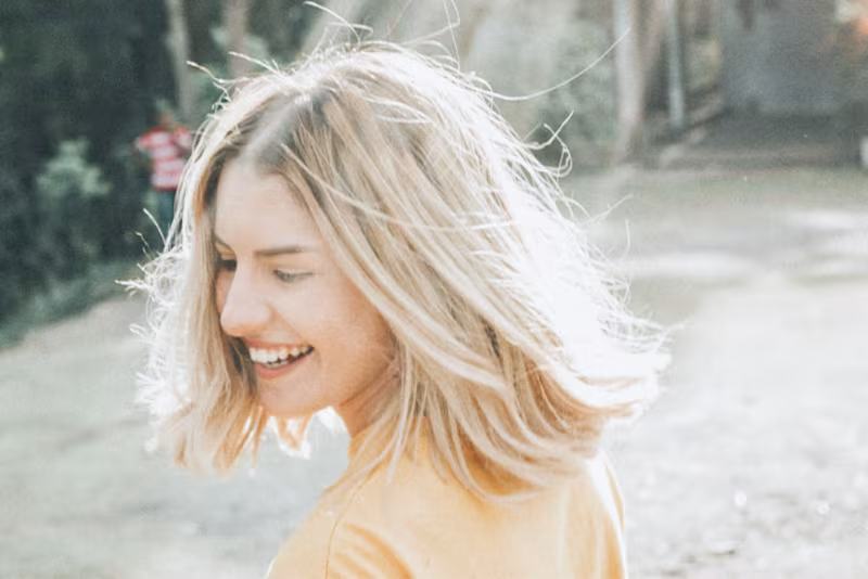 Lachende vrouw met wapperend blond haar kijkt over haar schouder, buiten in de zon